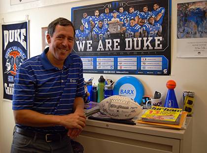 John Caccavale poses in his office, which is adorned with Duke posters and memorabilia, including a football autographed by members of the 2012 Duke football team. Caccavale is the 2013 honorary employee captain for the Employee Kickoff Celebration game.