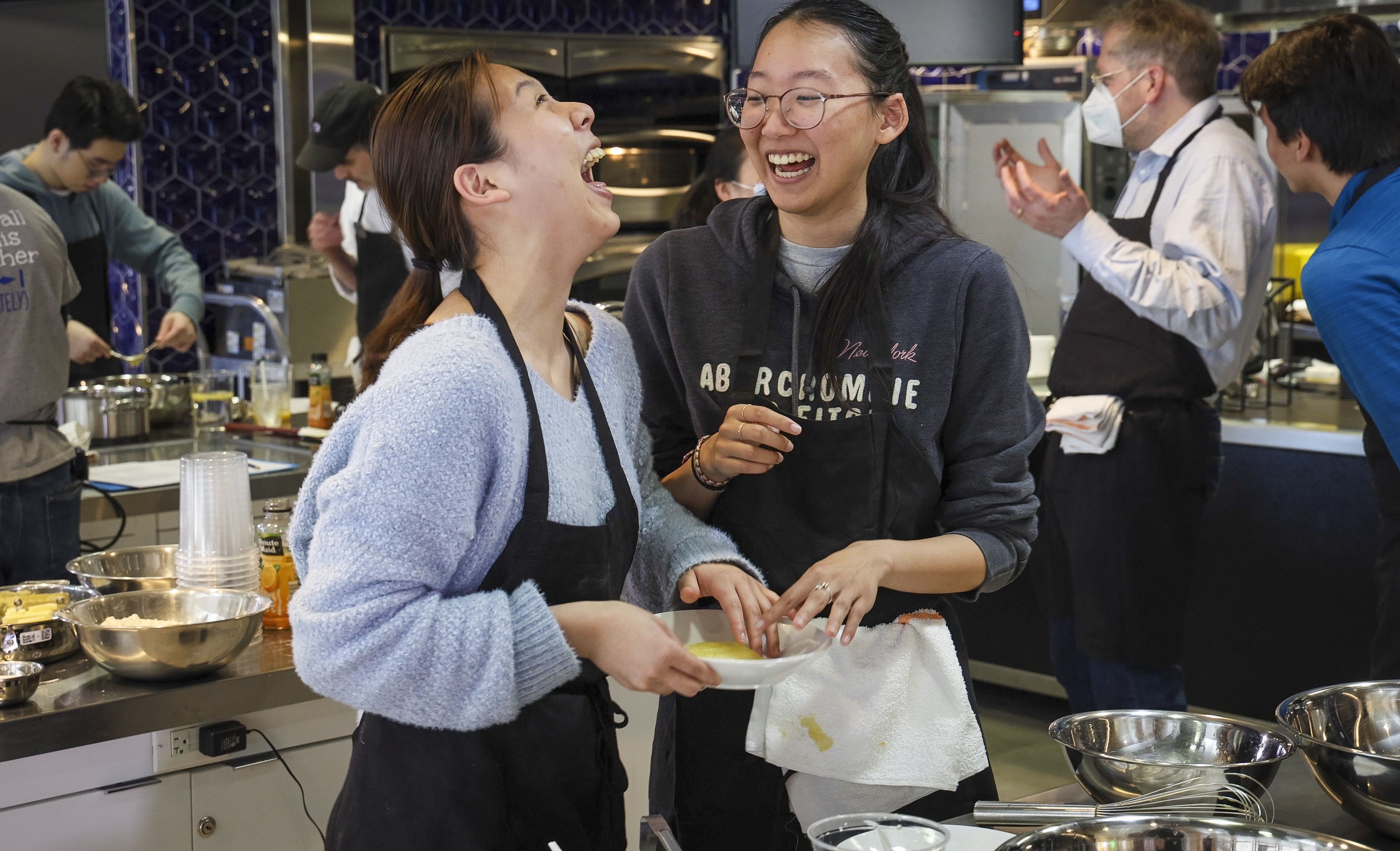 Two students laugh while cooking in a busy industrial kitchen