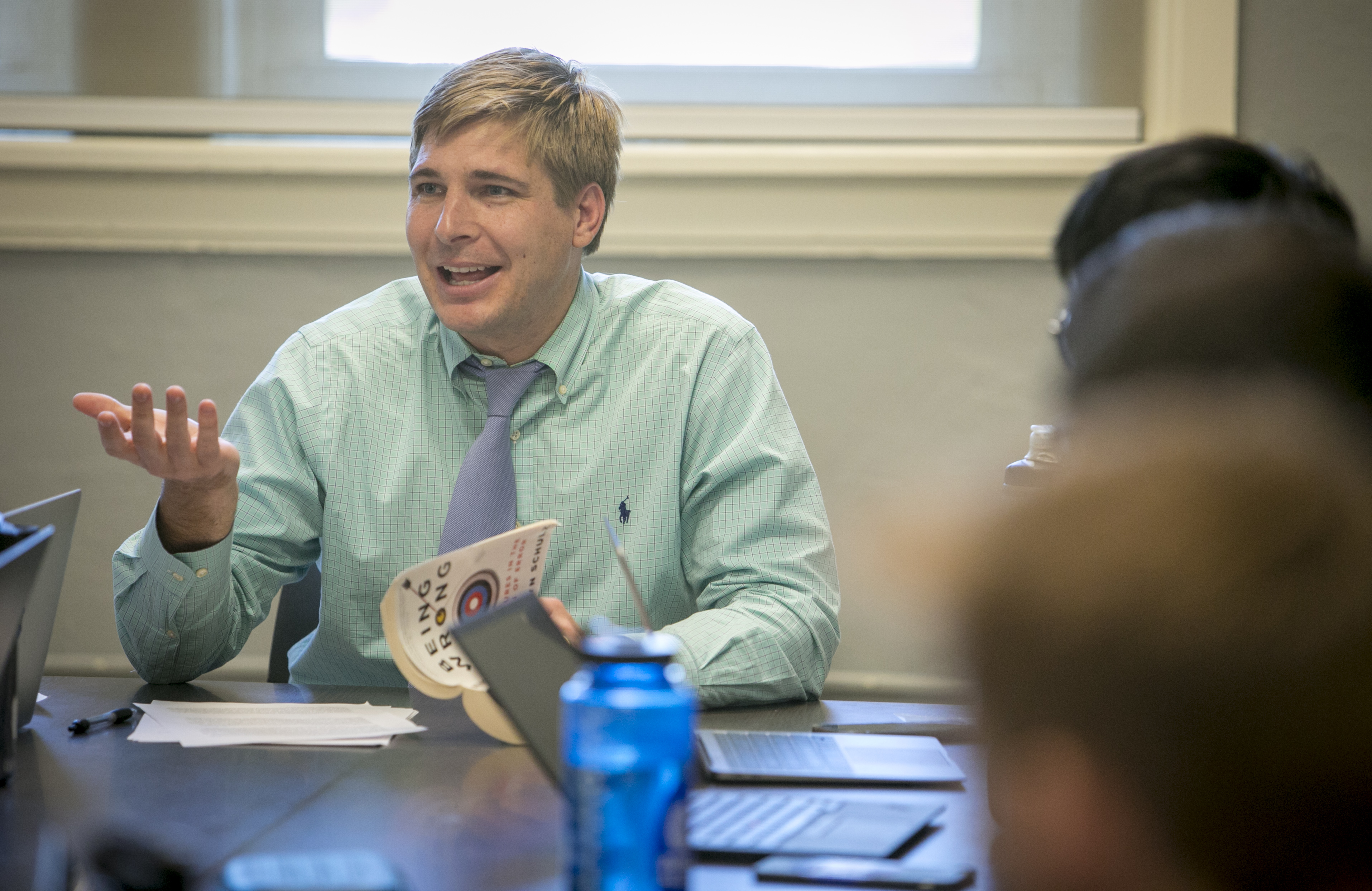 John Rose smiles and gestures to his class. Student heads are blurred in the foreground.