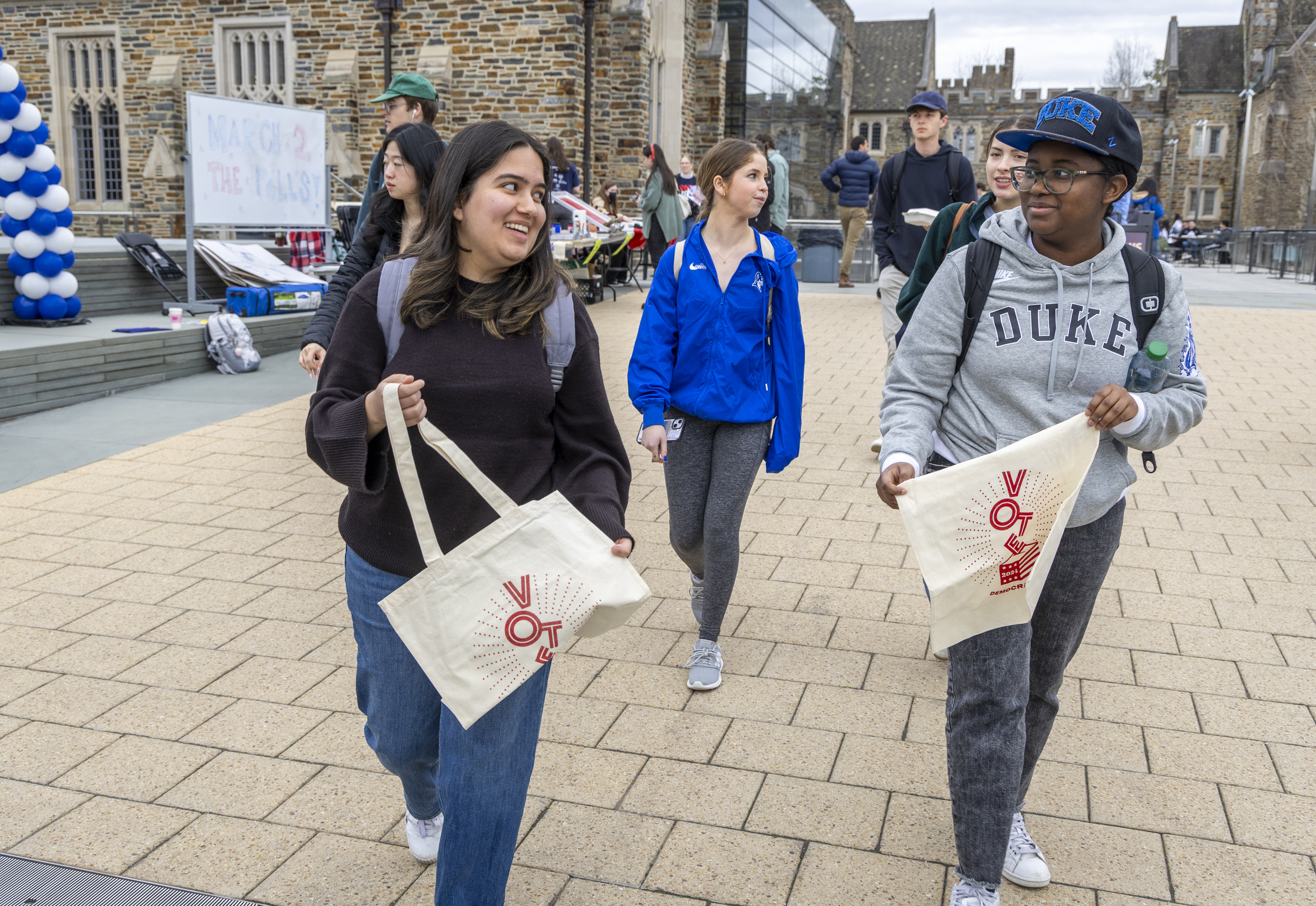 Students walk along Brodhead Plaza smiling and talking while holding tote bags with a red "Vote" message.