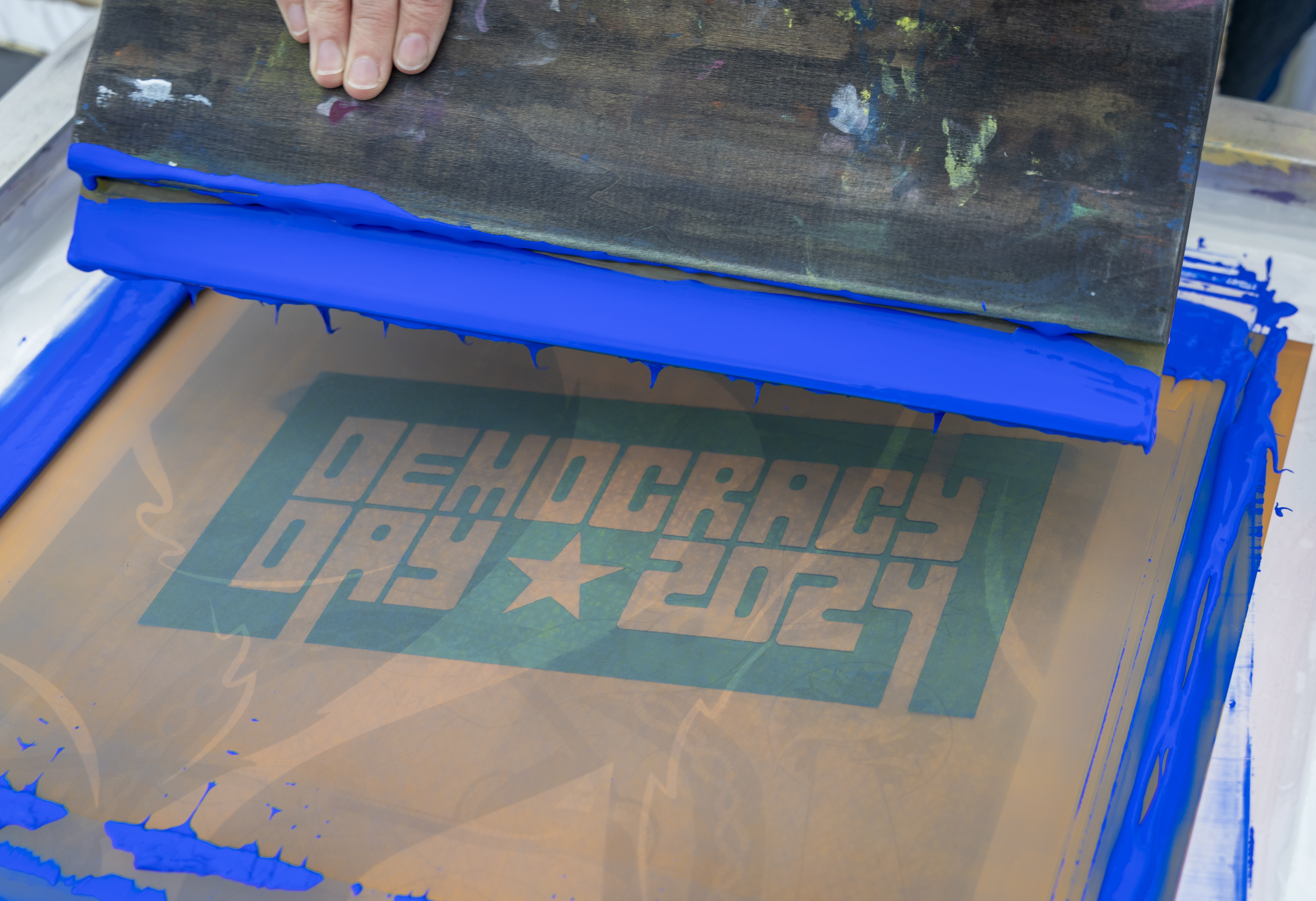 A hand holds a blade covered in bright blue paint, that has been dragged over a screen with the text, "Democracy Day" a star and then "2024"