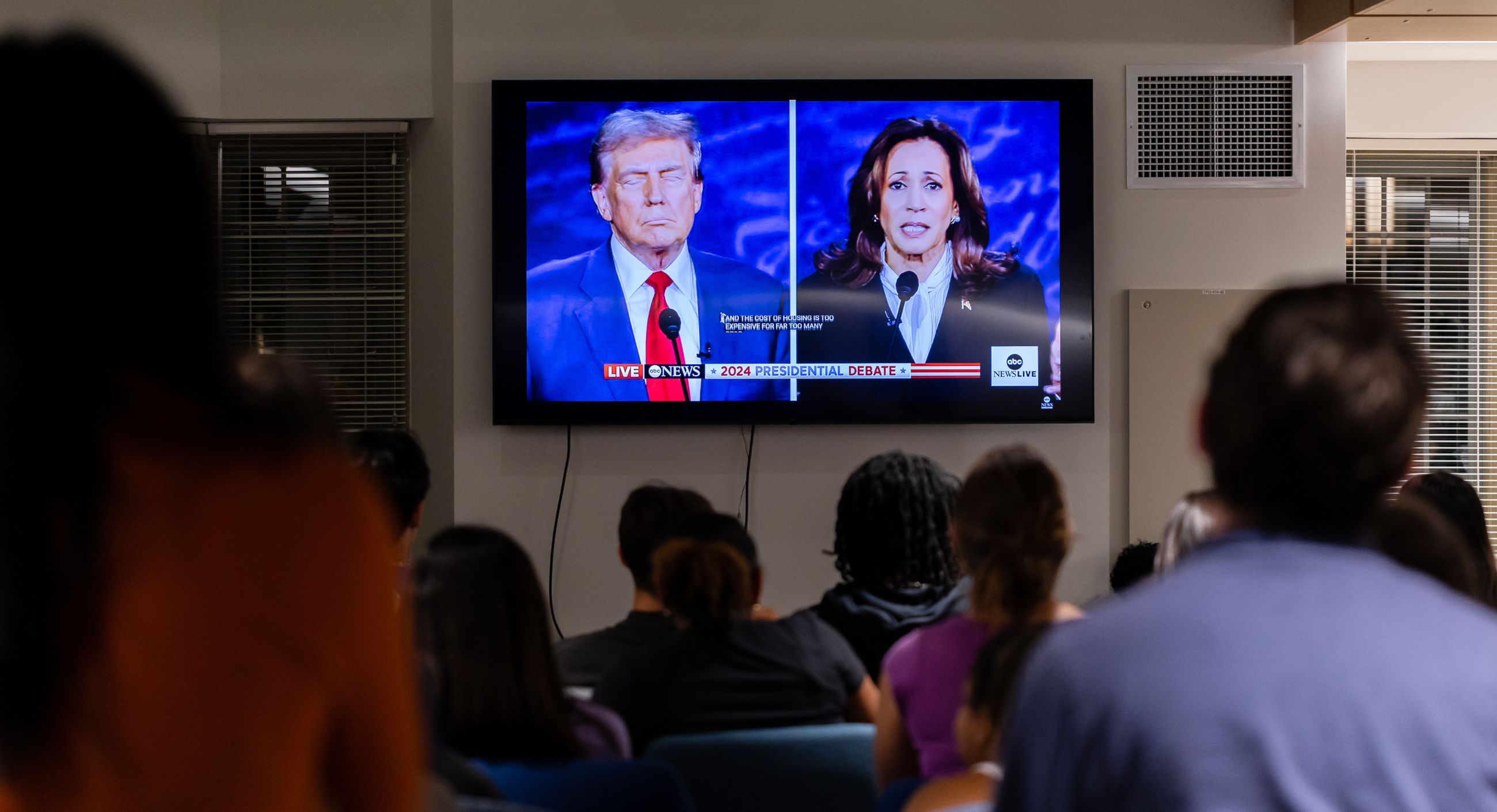 Donald Trump and Kamala Harris are shown on a TV screen during their presidential debate. Blurred student sit in the foreground with their backs to the camera.