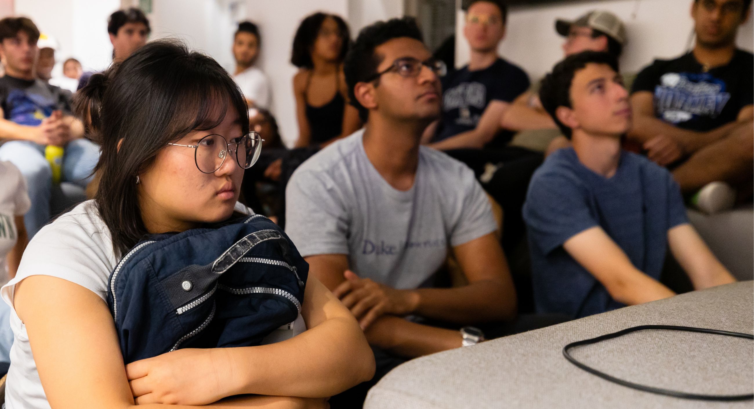 A student hold her backpack to her chest, looking concerned and she and others watch TV