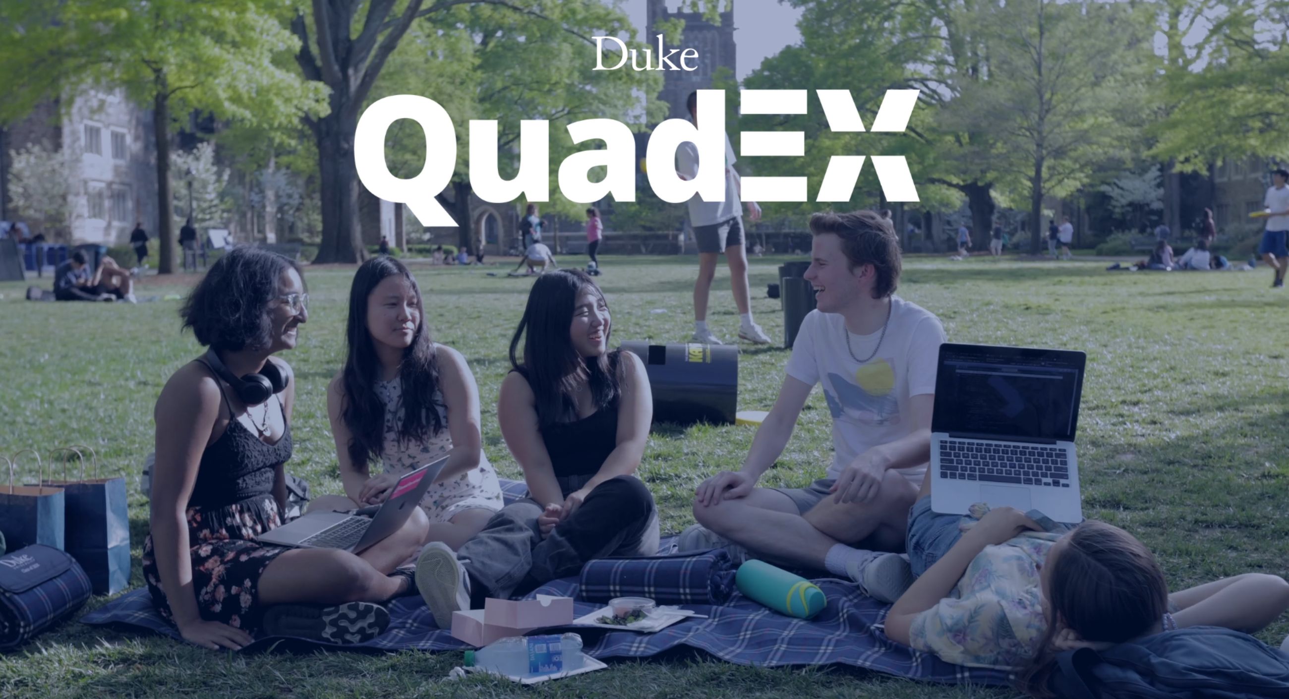 Students sit on the grass with laptops and food. The caption "Duke QuadEx" is in the upper third