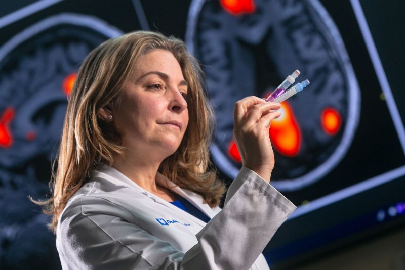 Heather Whitson compares the contents of two vials. In the background, large images of brain scans showing red highlighted areas.