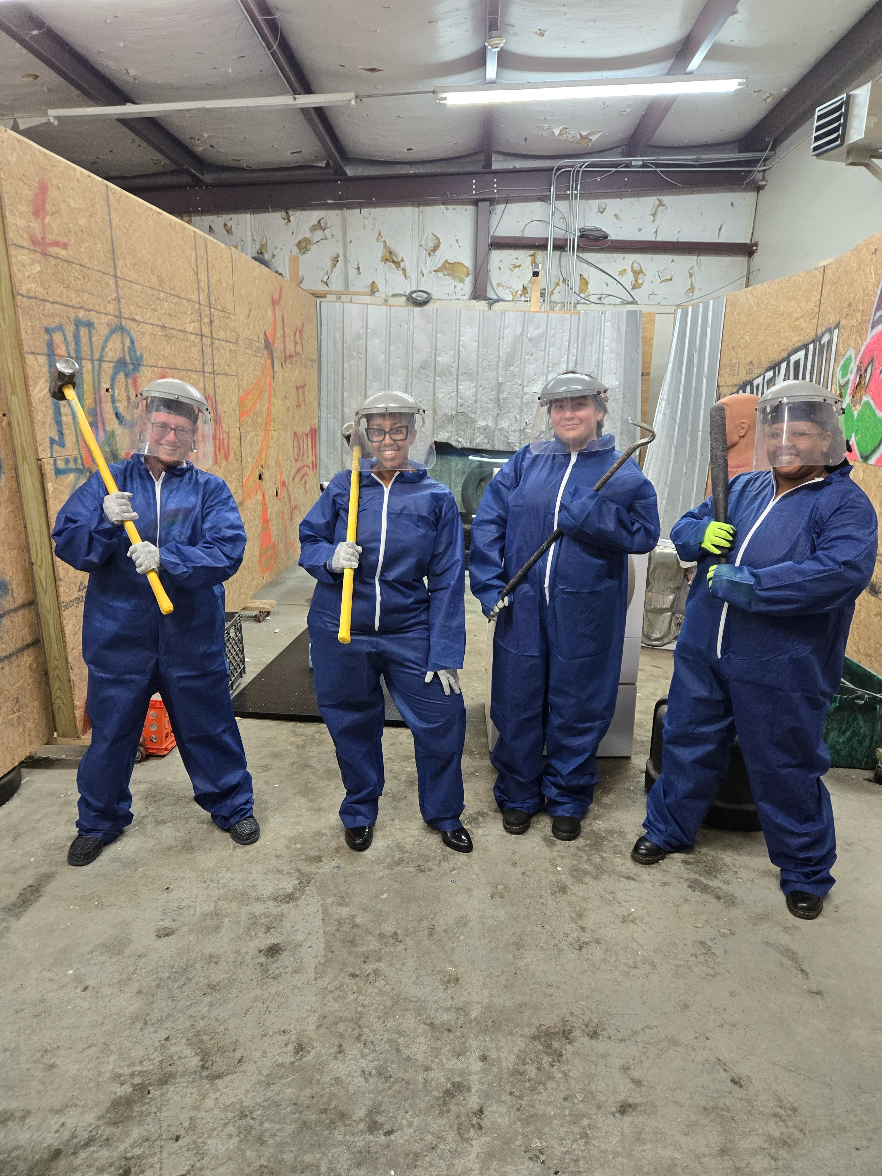 Four people stand in a "rage room," wearing coveralls and holding sledgehammers