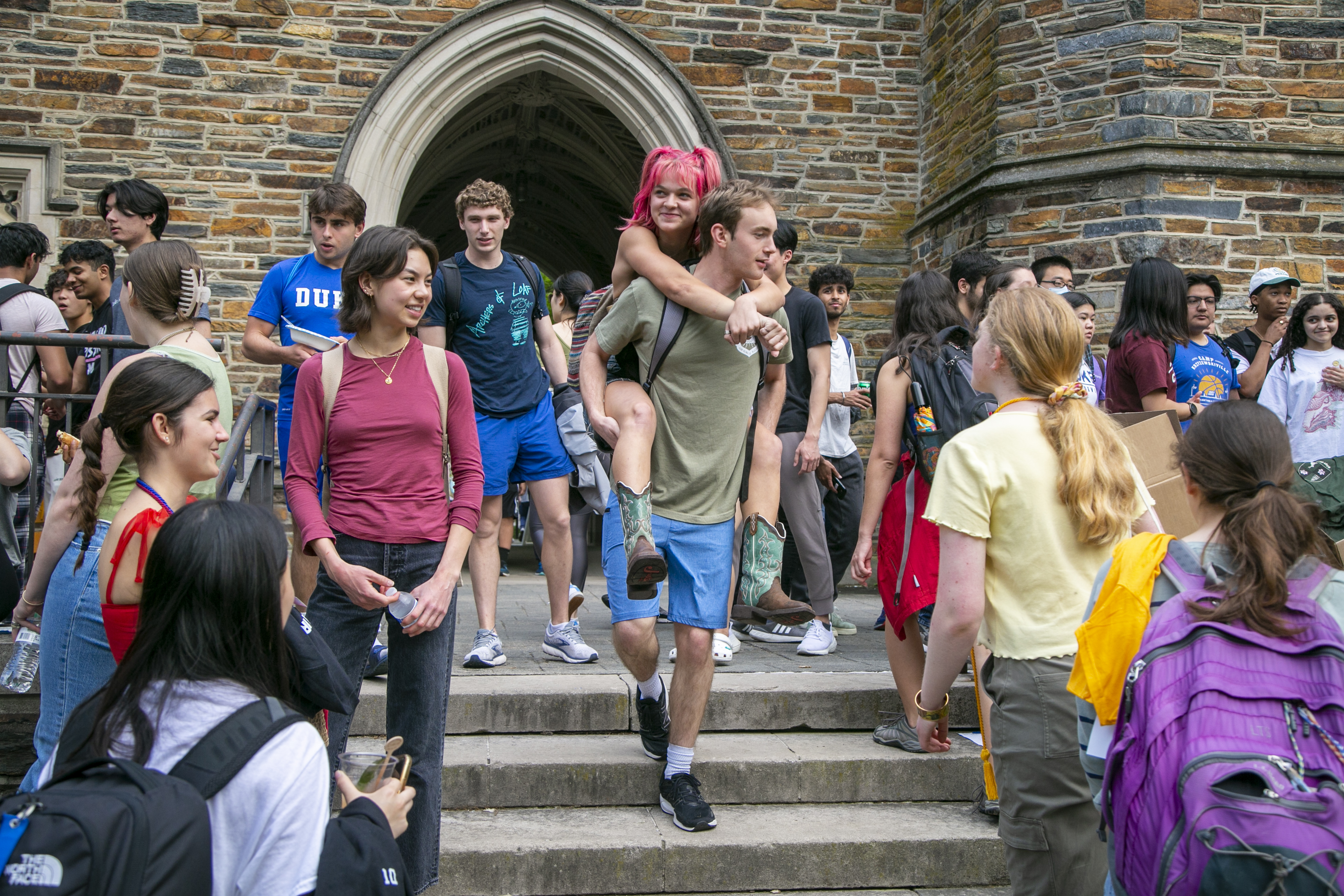 Students gather around and enter under the Kilgo Quad stone arch. In the center a student with bright red hair smiles, riding piggyback on a peer.