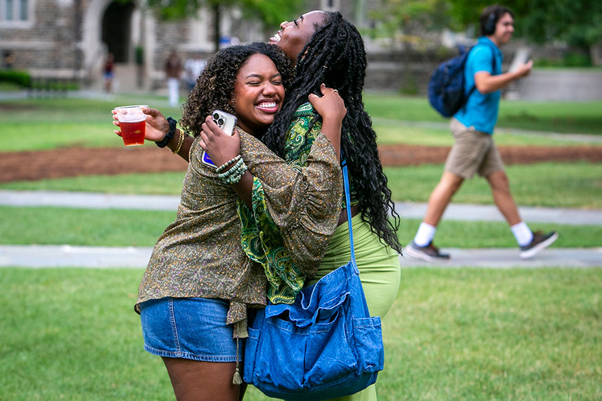 Two friends holding stuff embrace on the quad