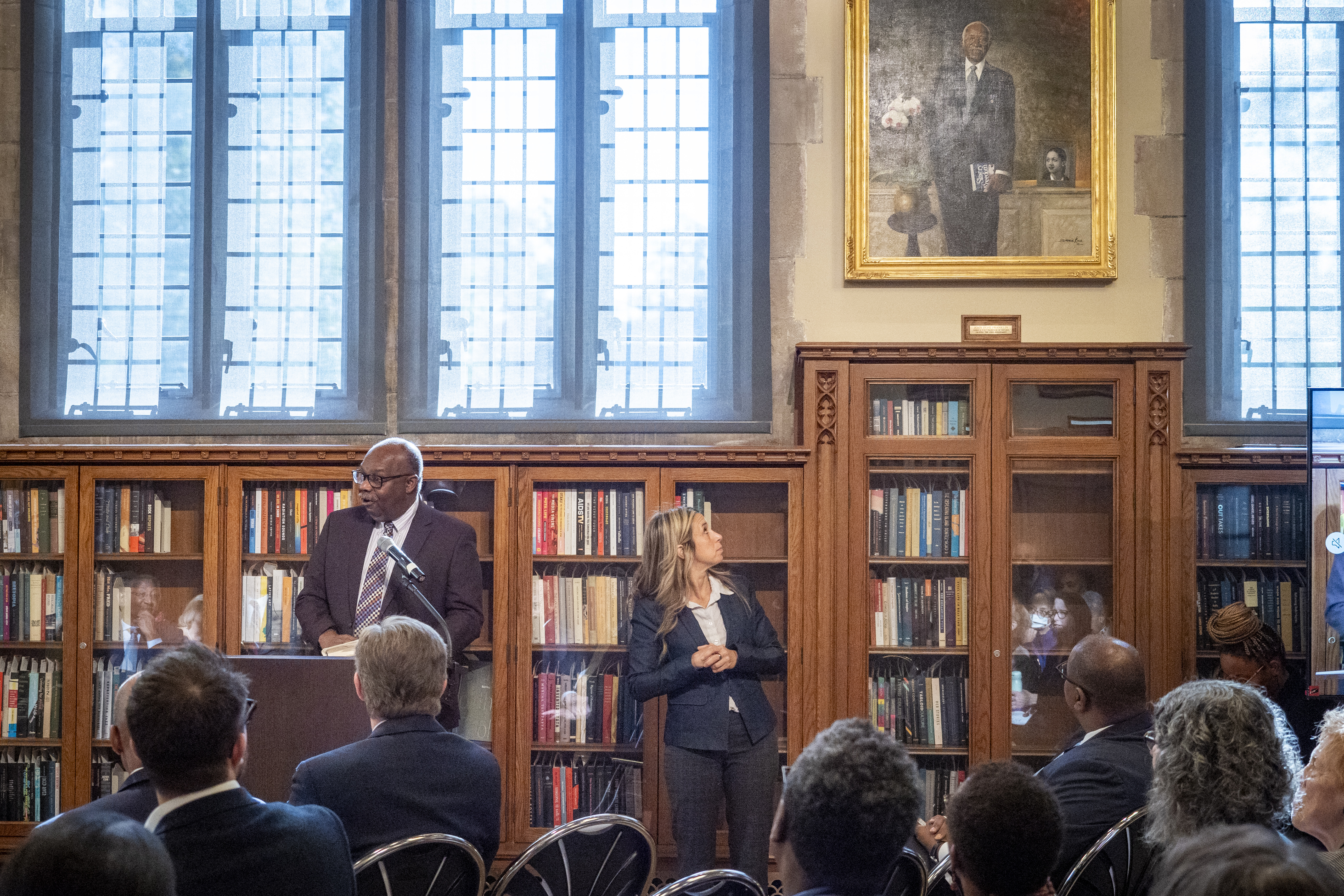 Dr. Kerry Haynie, speaks from a podium. He stands in front of a seated audience and behind him are the bookshelves and large windows of the Gothic Reading Room. To his right a sign language interpreter looks up and behind at a grand portrait of Dr. John Hope Franklin.
