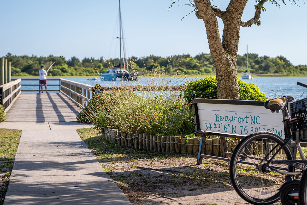 A picturesque photo of a bench painted with "Beaufort, NC" with a bicycle next to it and a pier in the background.