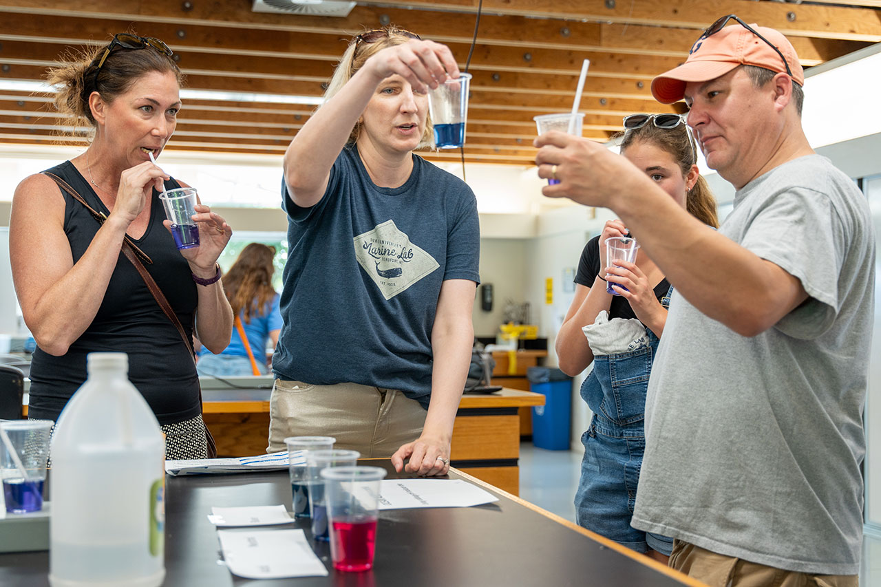Dana Hunt holds a cup of blue liquid while visitors blow through straws into cups demonstrating how carbon is captured making the liquid acidic.
