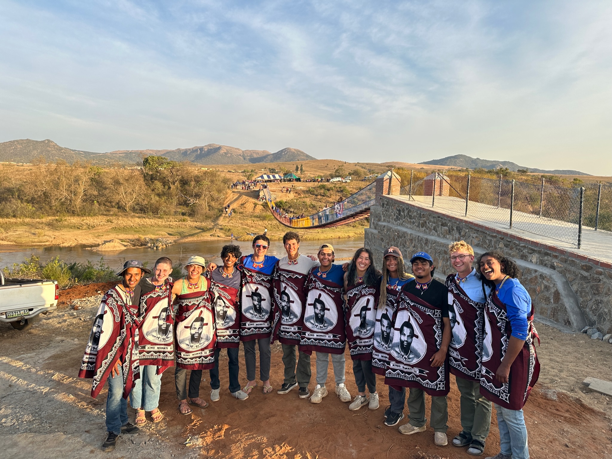 DukeEngage Eswatini students stand in front of the completed bridge on the day of the celebration. From left to right: Harsh Mathur, Emily Hallock, Maya Reeves, Nikhil Arayath, Kyle Abrahm, Palo Silva, Nimaye Garodia, Jackie Ong, Keely Blahauvietz, Diego Ayala, Harrison Kendall, and Anya Dias-Hawkins. Not pictured is Site Coordinator Syd Levy, who took this photo. The students are wearing gifts from community members, beaded necklaces and garments depicting Mswati III, King of Eswatini. 