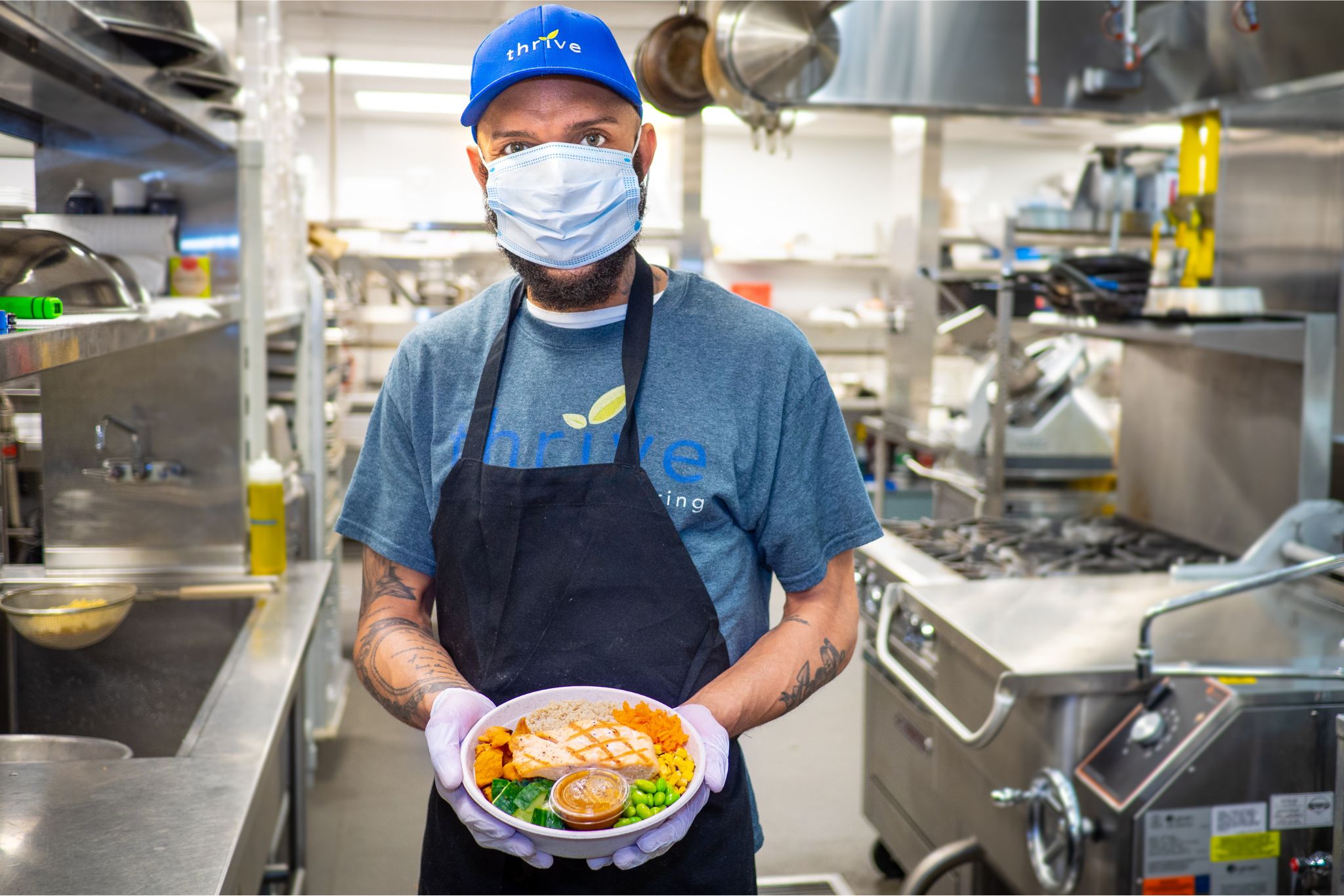 Lucas Joseph Ramsey, cook for Thrive Catering, stands in the kitchen holding out a rice bowl with beautifully grilled chicken and vegetables.