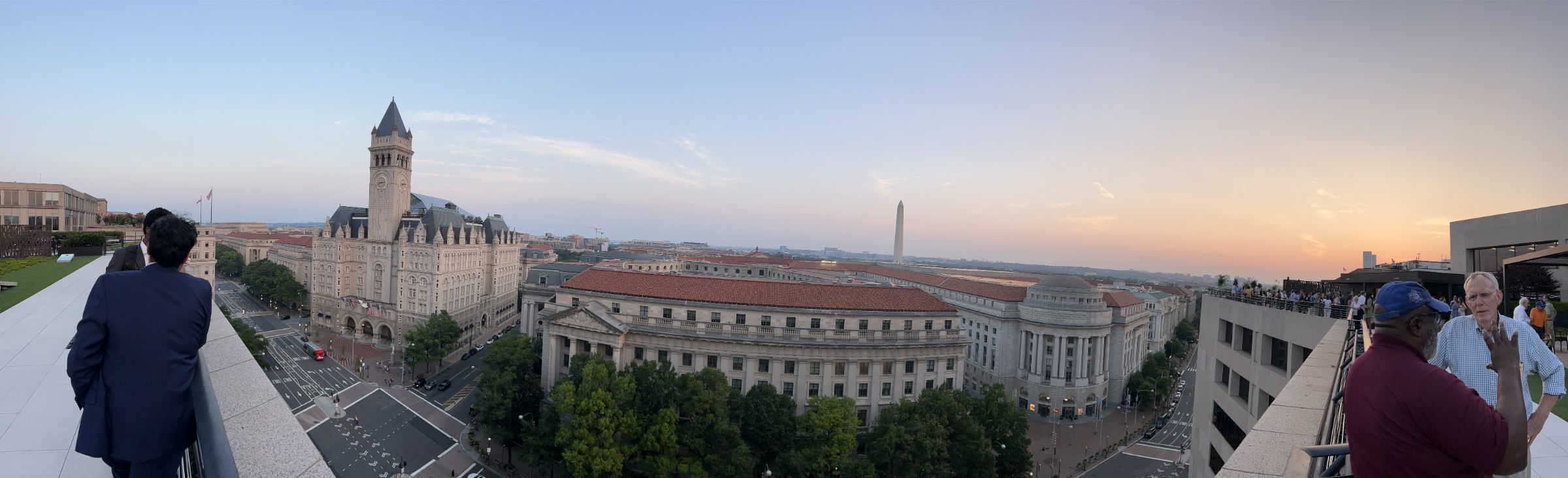 Panoramic view of downtown Washington, DC. To the left and right people are leaning on the balcony wall talking.  In the center are prominent DC building such as the Old Post Office Pavilion the and Washington monument