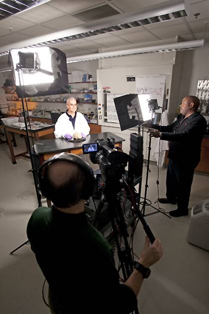 TV lights and a camera surround Len White standing at a table in a white lab coat, a human brain sits on the table in front of him.