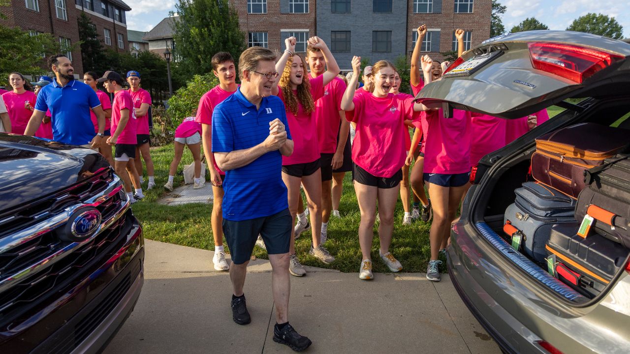 President Vincent Price, in a blue shirt with a Duke "D", steps toward the back of a car as the lift rises to show suitcases. Behind him are cheering students in pink shirts, waiting to help move a new student into their dorm.