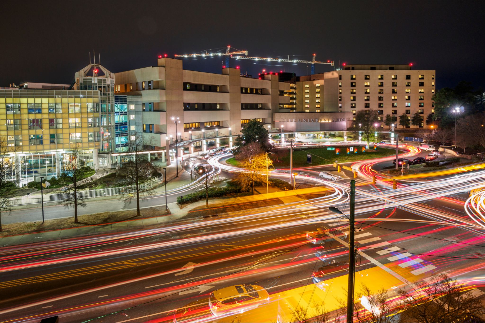 At night, in front of the main entrance of the Duke University Hospital, a long photo exposure time shows the red and white light trails of cars that have passed. A single white mini-van is in the foreground.