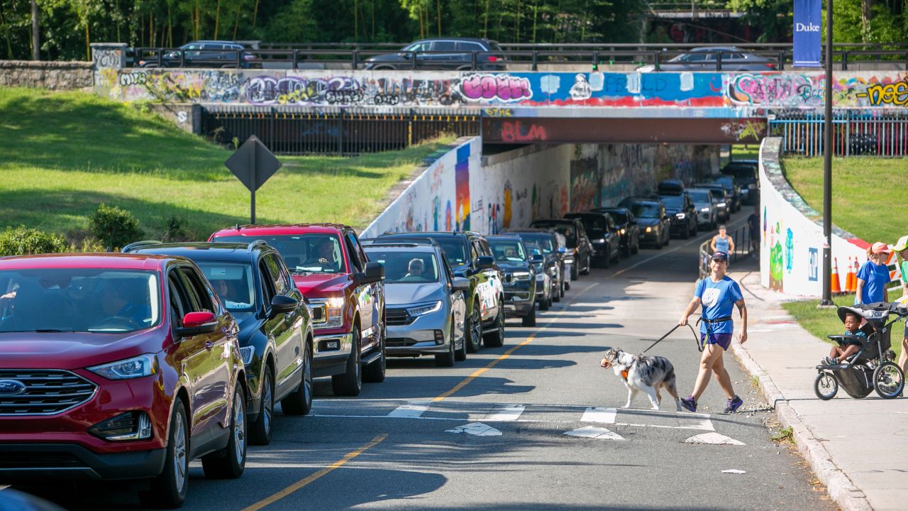 A line of cars emerge from a graffiti covered overpass. Pedestrians with a dog and baby in a stroller walk between cars at a cross-walk.