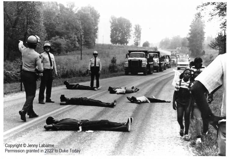 Six Black protesters lie in the road so trucks cannot pass, while white police officers stand over them deciding what to do.