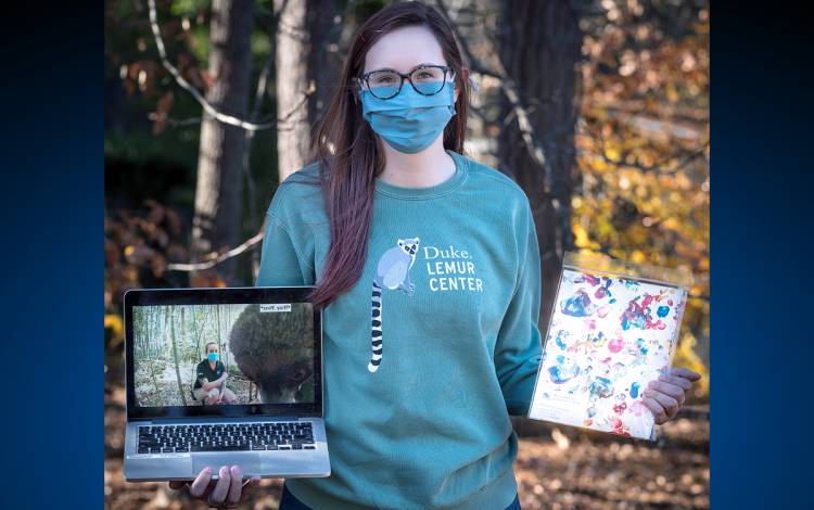 Among the gifts available from the Duke Lemur Center are virtual experiences, apparel and even paintings by the lemurs themselves. Photo courtesy of Duke Lemur Center. A person holding a laptop and painting.