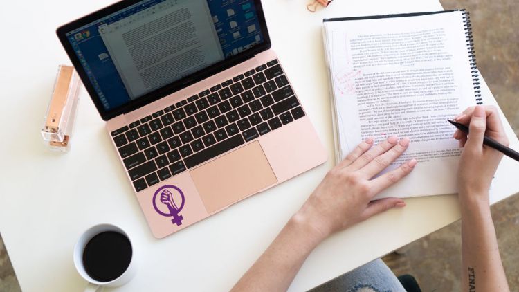 View of white table from above. Cup of coffee in lower left. rose-colored laptop with women's empowerment symbol decal in center, hands holding manuscript and pen making updates, in lower right.