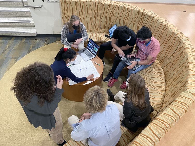 A group of students with computers sits in a lounge. In the middle, a woman draws image on a large sheet of paper on a coffee table. 