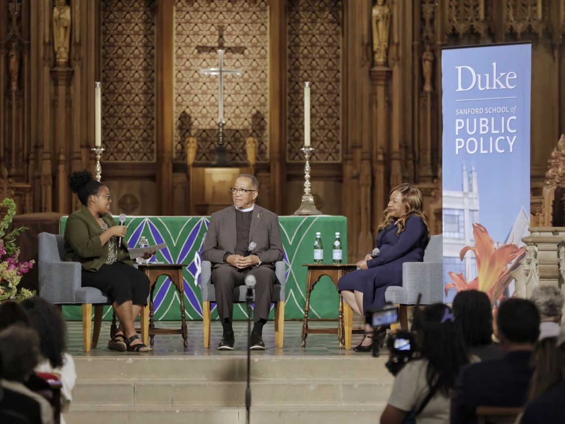 Three people sit in the front of the Duke Chapel next to a banner that reads,