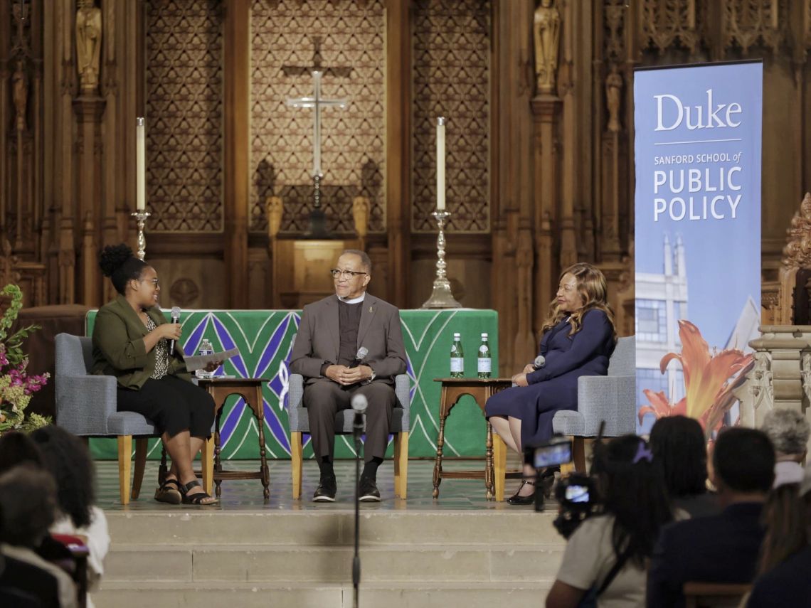 Three people sit in the front of the Duke Chapel next to a banner that reads, 