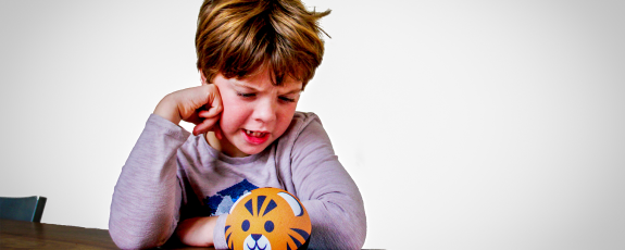 An 8-year-old boy at a table, his head resting on his fist, appears to be having angry words with a kid-friendly Alexa device that looks like a cat's head.