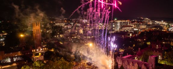 An aerial view of fireworks bursting over West Campus with Duke Chapel and Duke Hospital in the background