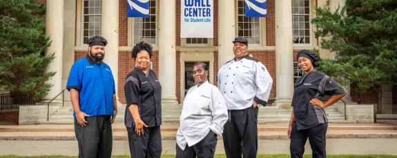 Five people in food service uniforms pose for the camera on the lawn in front of a large university building, made of brick with columns of sandstone. Banners celebrating Duke's Centennial and the Wall Center dedication hang between the columns.
