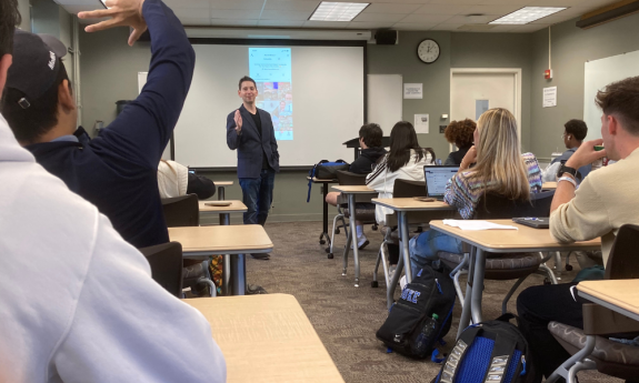 Aaron Dinin stands at the front of a classroom with TikTok on a screen behind him.  In the foreground a student is raising their hand.