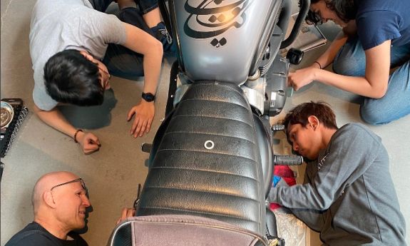 Overhead view of three students and instructor sitting or lying next to a motorcycle they are working on.