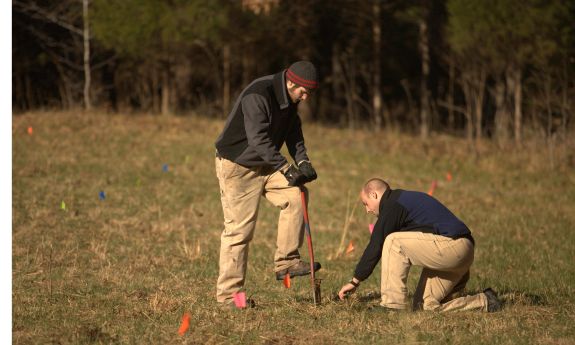 Two men in khaki pants and dark tops work together to plant a small tree.