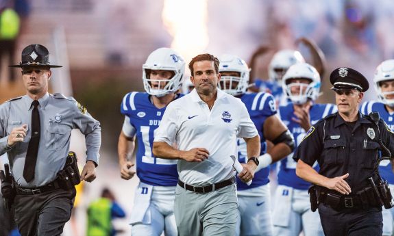 Duke head football coach Manny Diaz runs onto the field, flanked by police officers and followed by players in uniform 