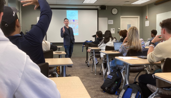 Aaron Dinin stands at the front of a classroom with TikTok on a screen behind him.  In the foreground a student is raising their hand.