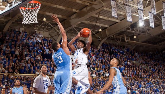 Duke's Justise Winslow leaps to dunk a basketball while UNC's Kennedy Meeks attempts to block the shot. Several players watch the play develop and hundred's of fans cheer in the background in Cameron Indoor Stadium