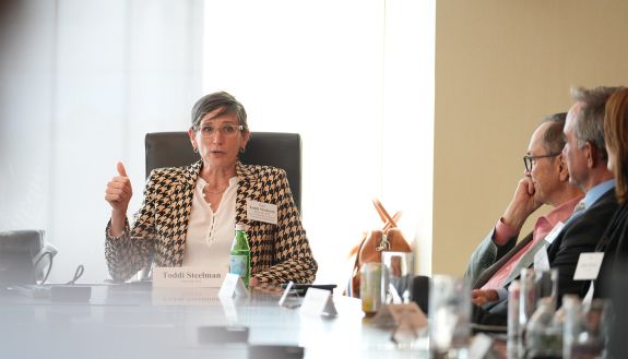 Vice President Toddi Steelman, wearing a houndstooth jacket, sits at the head of a conference table.