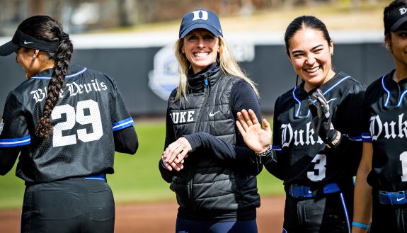 A Duke instructor stands on the sidelines of a softball field among three Duke softball players before a softball game