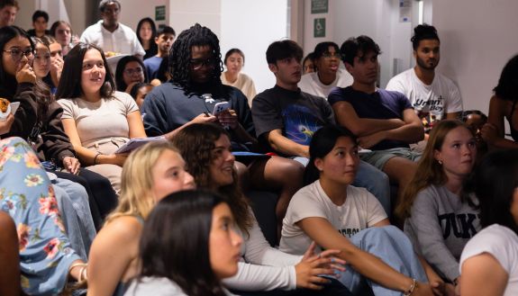 Students in a dorm, some looking shocked, watch a program on TV.