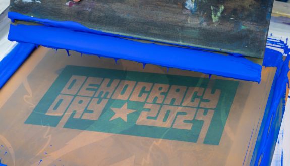 A hand holds a paint scraper over a screen print screen with blue paint dripping. the screen says "Democracy Day 2024"