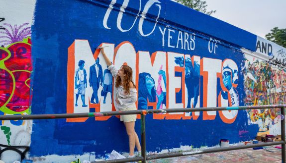 A student leans up against a wall to add paint to the letter "M" in the nearly complete word "MOMENTS". Images of people and events surround and intersect with the letters.