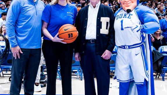 Four people on a basketball court with one holding a ball and on the right is the Blue Devil