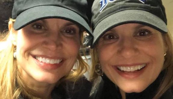 Twins wearing baseball hats. On left UNC hat on right Duke hat