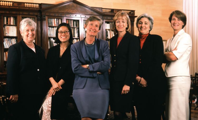 Six women stand in a row, smiling in front of a grand bookshelf