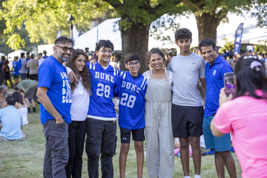 Seven people including two first-year students pose for a photo under large trees on the quad while someone takes their photo with a phone