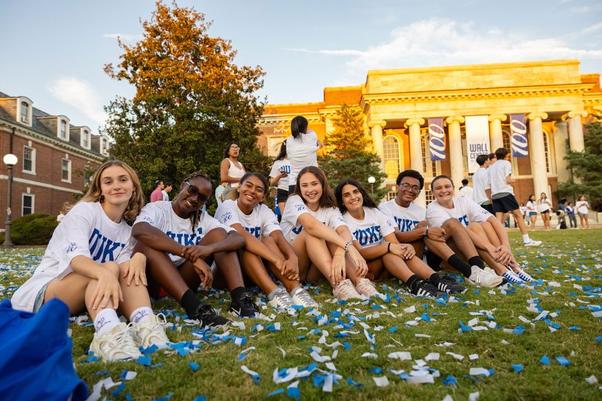 A group of seven students sitting in the grass among fallen confetti in front of the Wall Center on East Campus