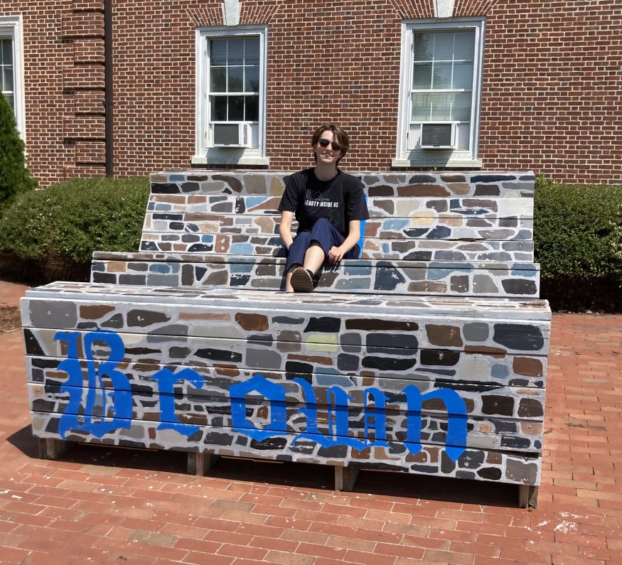 Meehan sitting on a large bench that is painted like Duke stone with "Brown" written in blue across the front
