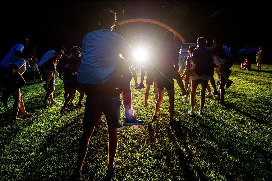 An orientation group giving piggy back rides in the dark with a car light illuminating the field