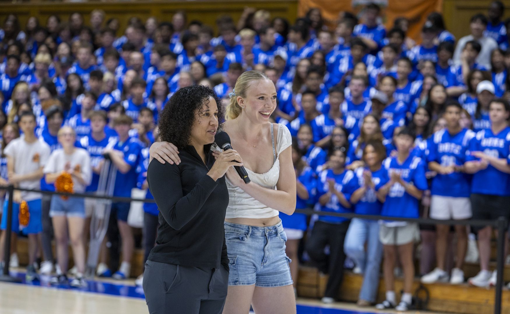 Basketball player Toby Fournier stands with her arm around Coach Lawson as she holds a microphone and addresses the first-year students and parents in Cameron Indoor stadium